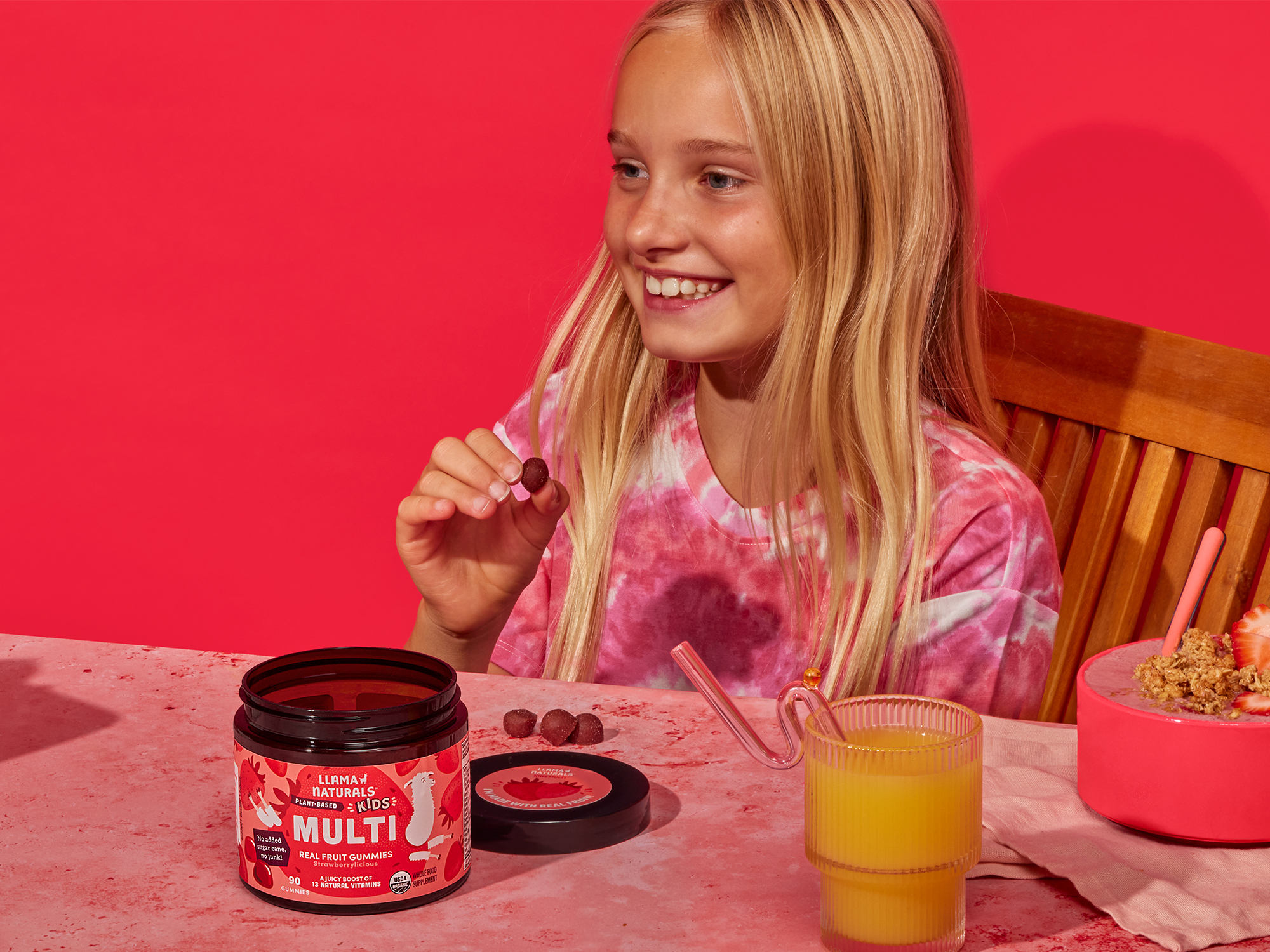 Smiling child holding a gummy, with juice and cereal on a red table.