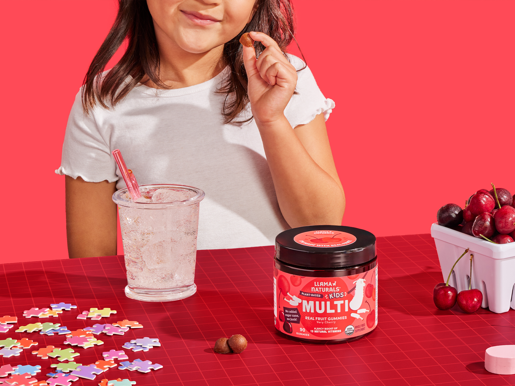 Child with gummies, drink, puzzle pieces, and cherries on a red table.