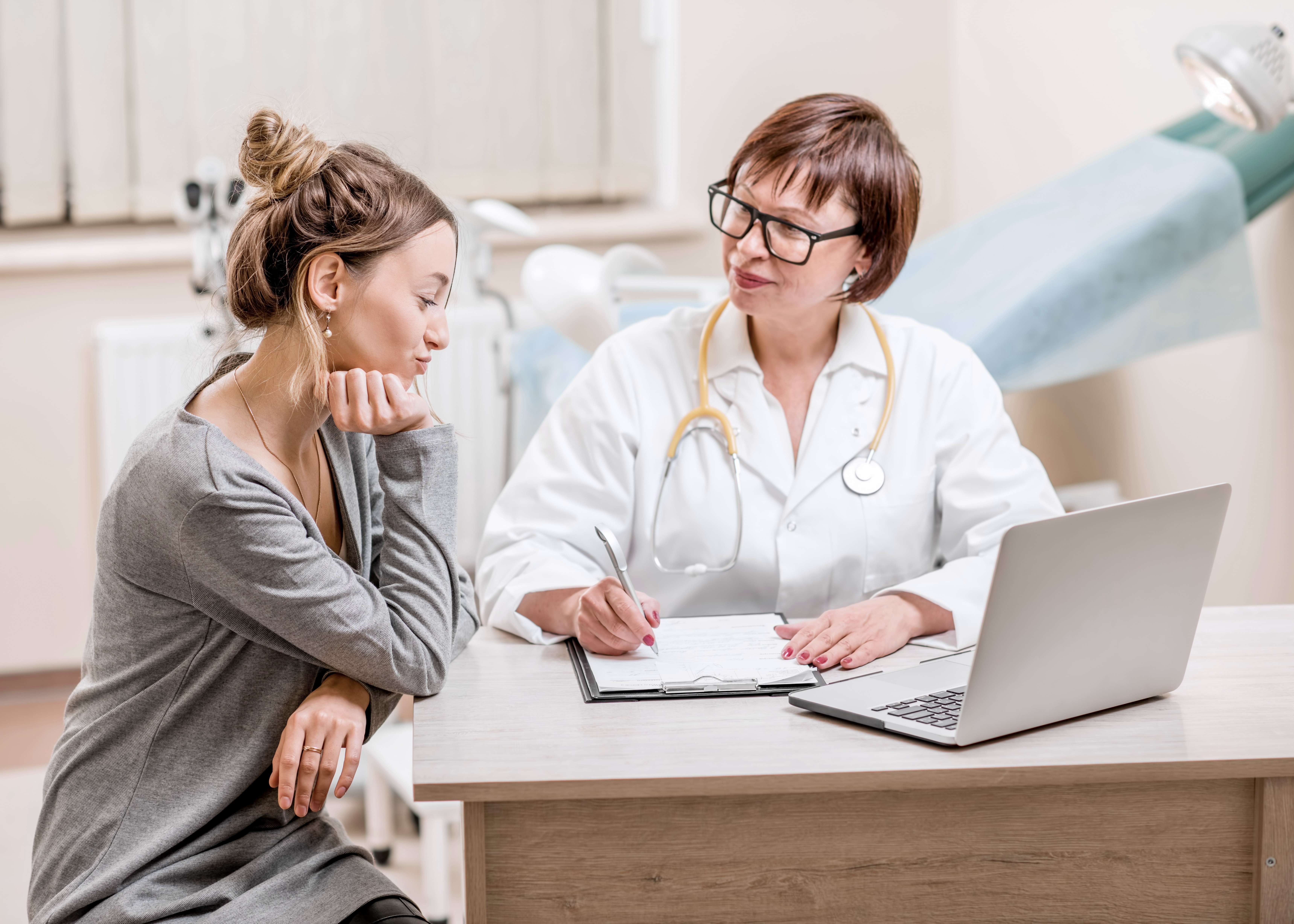 A female doctor in a white coat talks with a young female patient at a desk in her office.