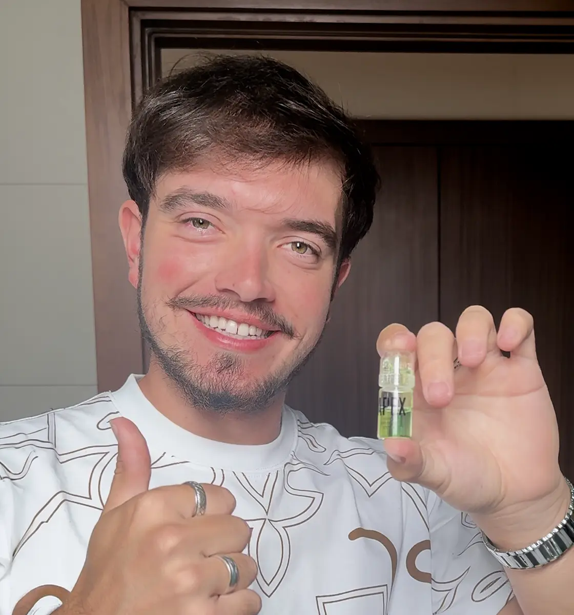 A smiling man with a mustache holds up a small vial and gives a thumbs-up sign.