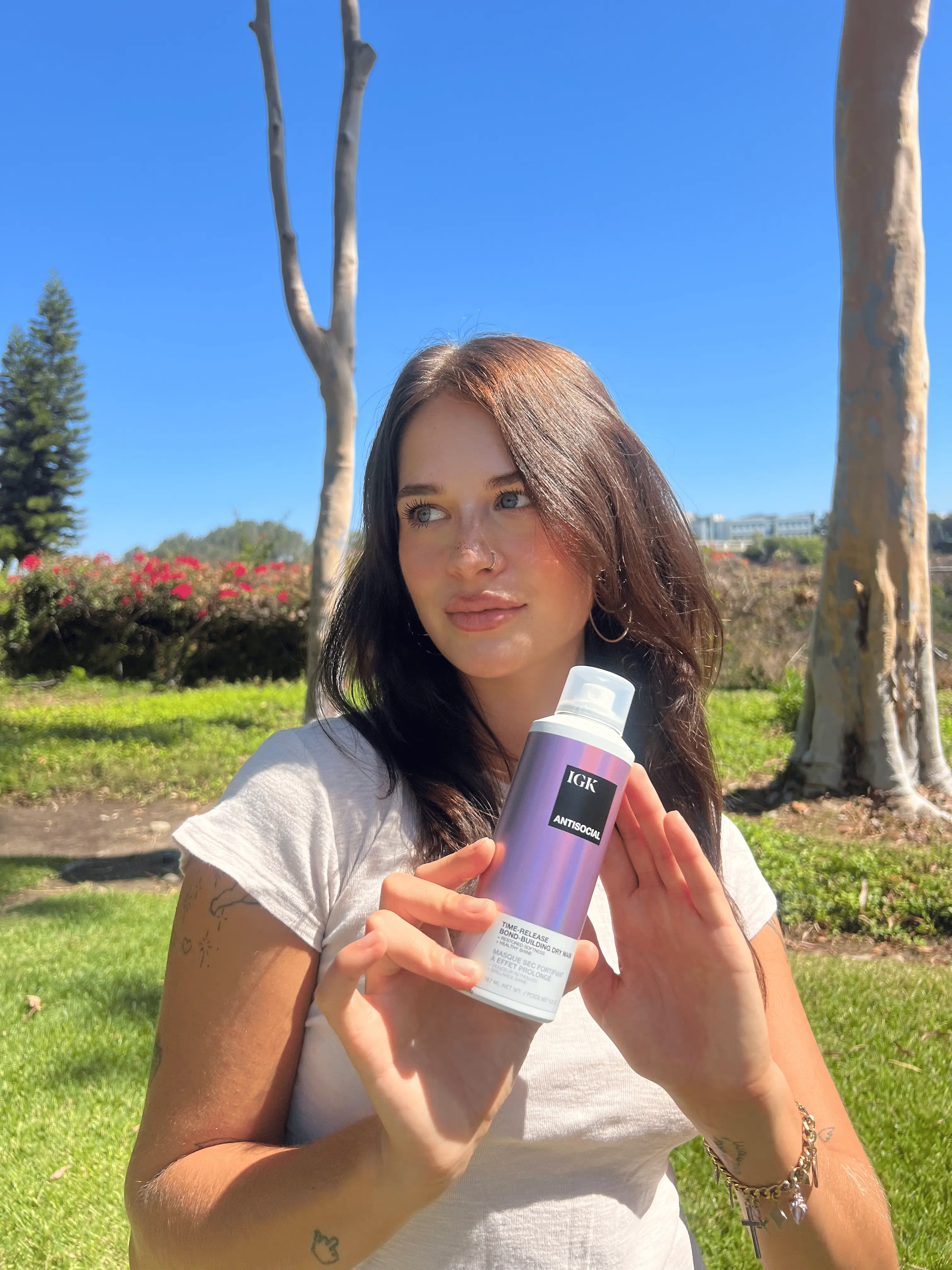A woman with long brown hair holds a can of IGK Antisocial hair product in a sunny park.