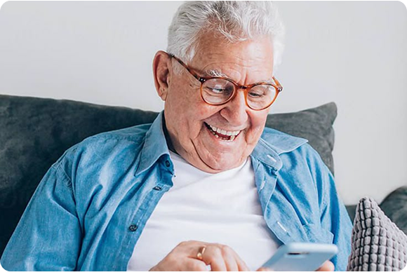 Elderly man smiling while using a smartphone on a couch.