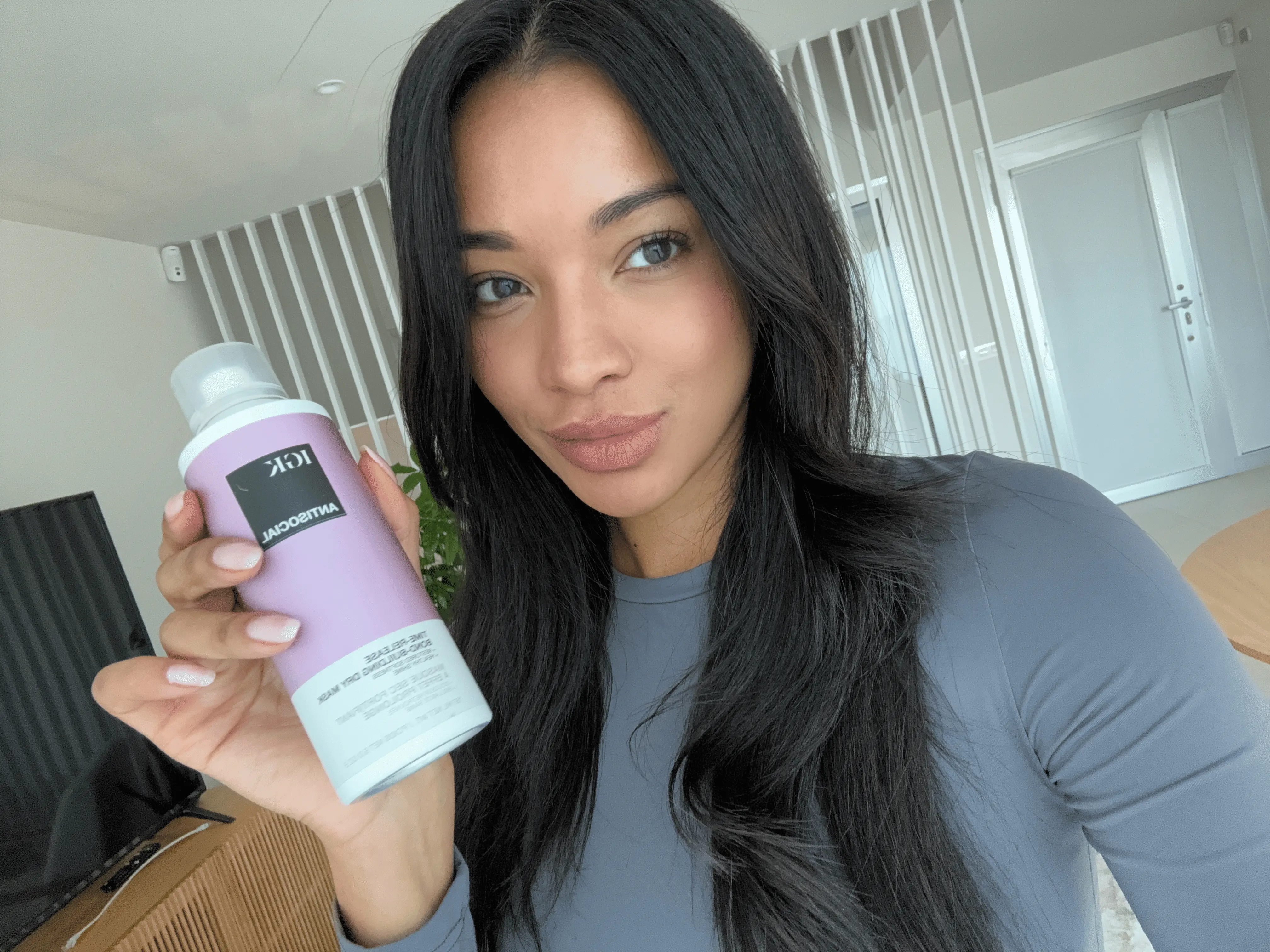 A woman with long dark hair takes a selfie while holding a purple can of IGK Antisocial hair product.