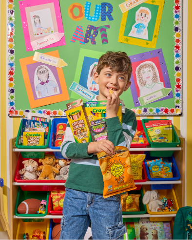 A smiling woman and a boy sit at a table covered with art supplies and snacks.