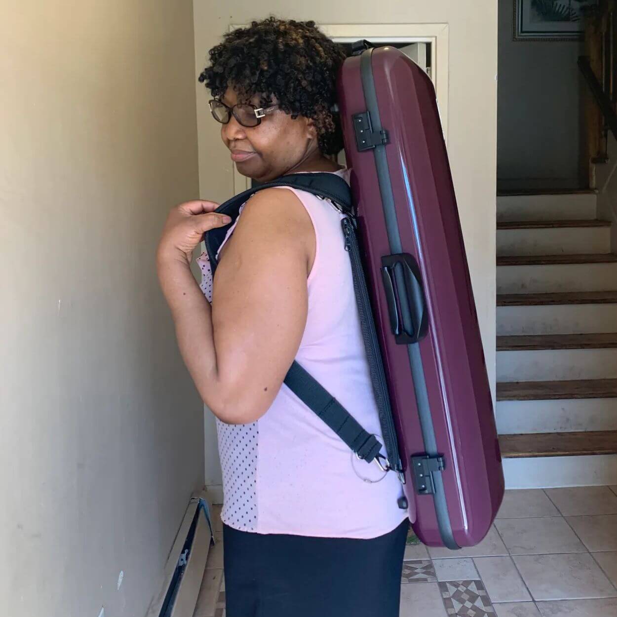 A woman with short curly hair and glasses carrying a large, purple hard-shell instrument case on her back.