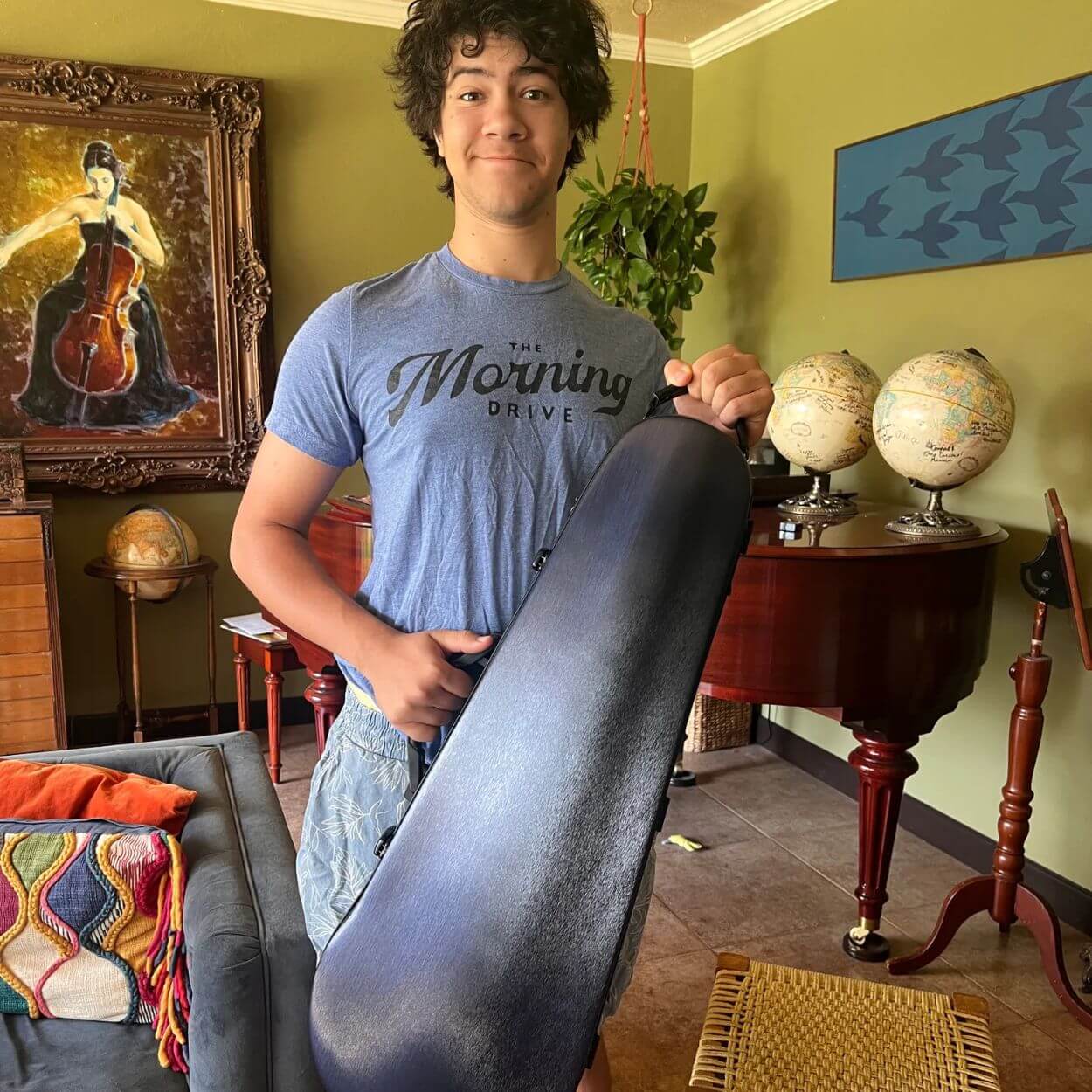 A young man with dark curly hair smiles while holding a string instrument case in a room.