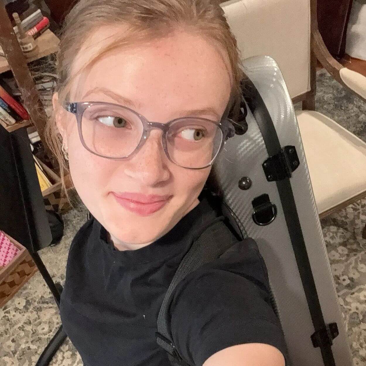 A young woman with glasses and a black t-shirt smiles while carrying an instrument case on her back.
