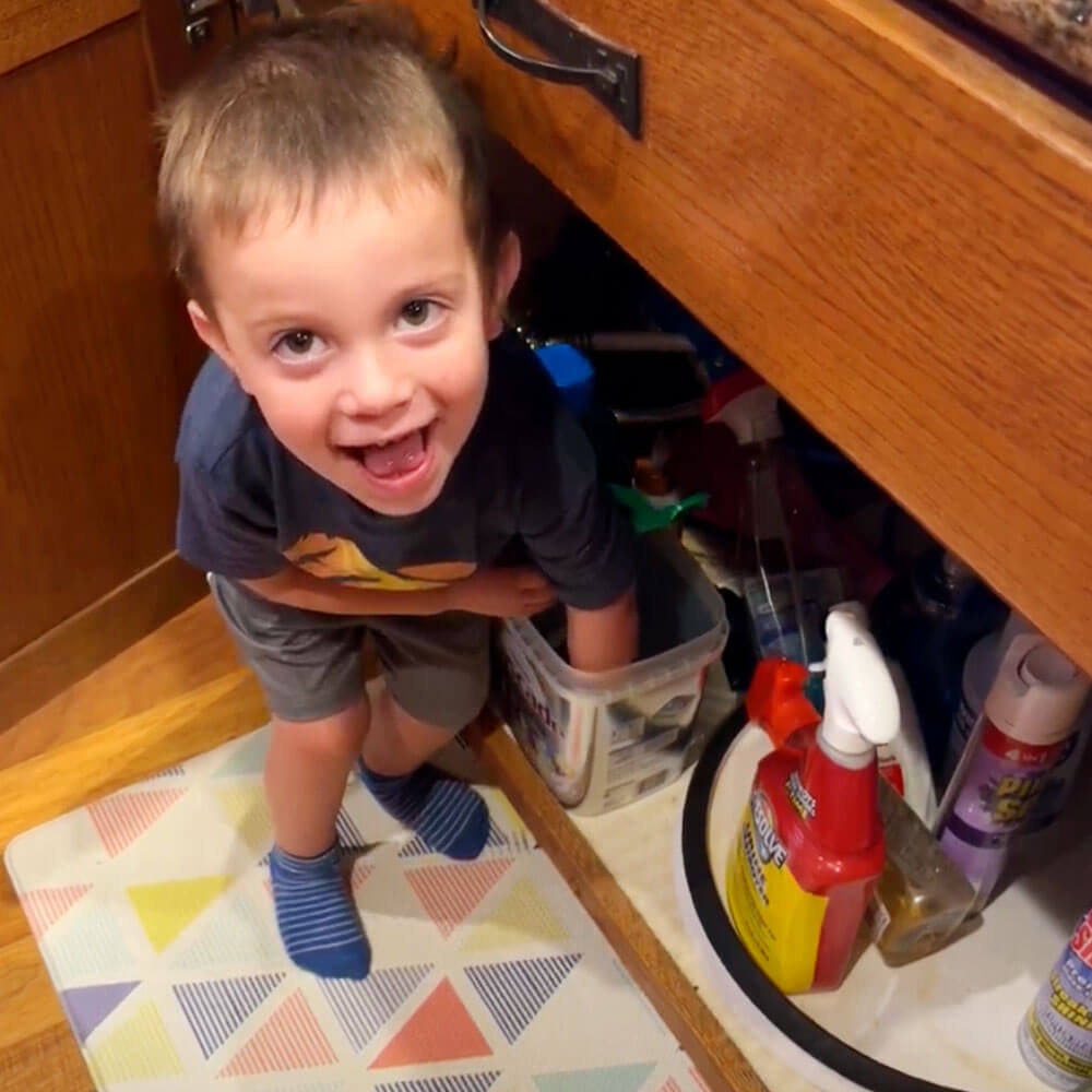 Child looking up, smiling, standing by an open cabinet with cleaning supplies inside.