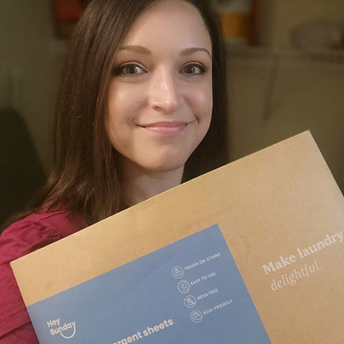 A smiling woman with brown hair holds a package of Hey Sunday laundry detergent sheets.