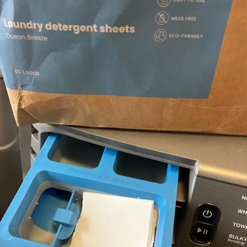 A laundry detergent sheet placed in the dispenser drawer of a washing machine, with the product packaging on top.