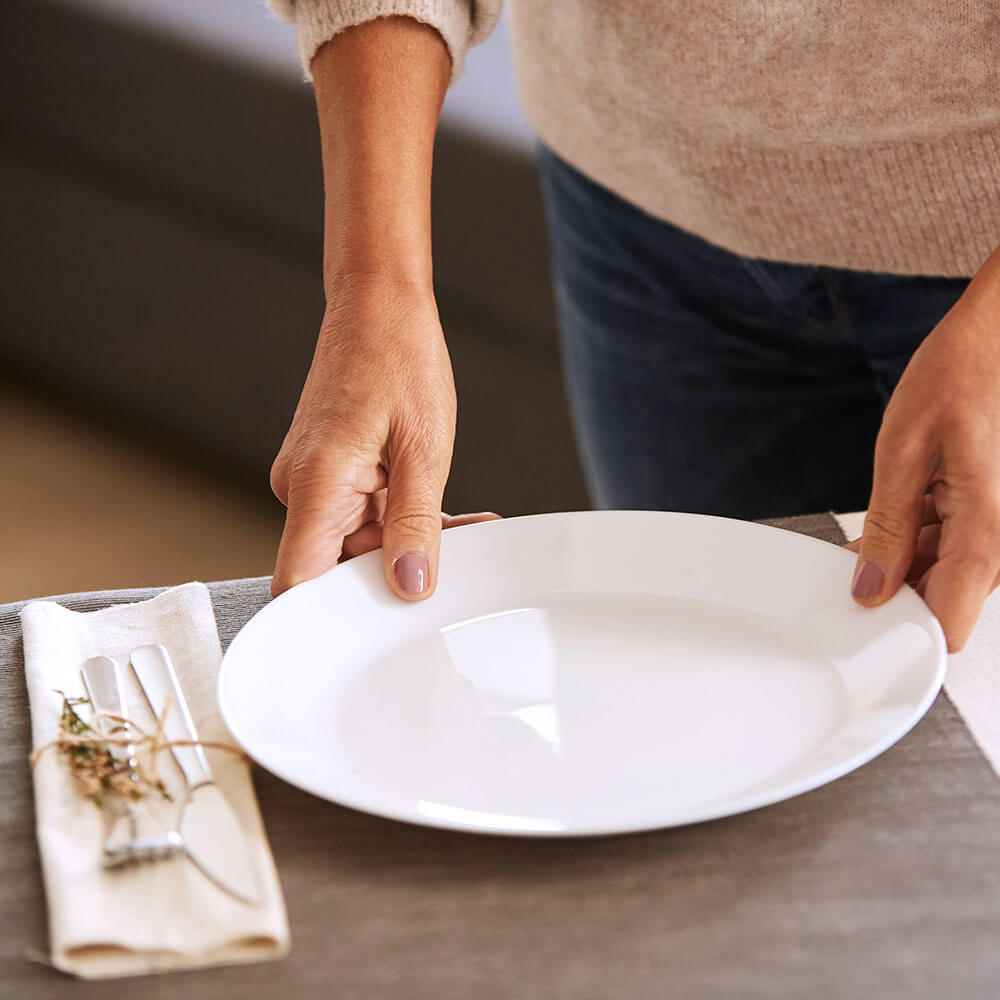 Person placing a white plate on a table; cutlery beside it.