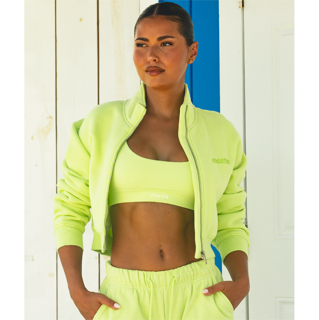 A woman with her hair pulled back wears a lime green sports bra and matching open cropped jacket.