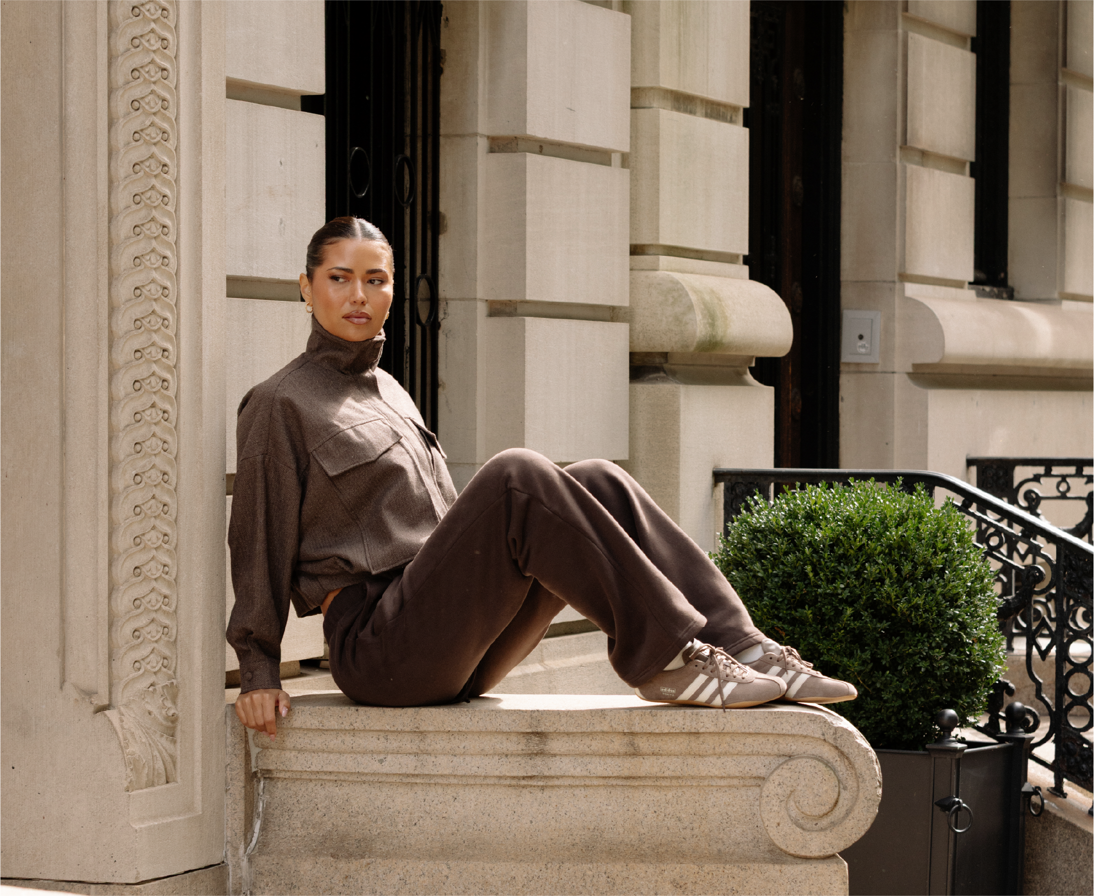 A woman in a brown outfit and sneakers sits on a stone ledge outside a building.