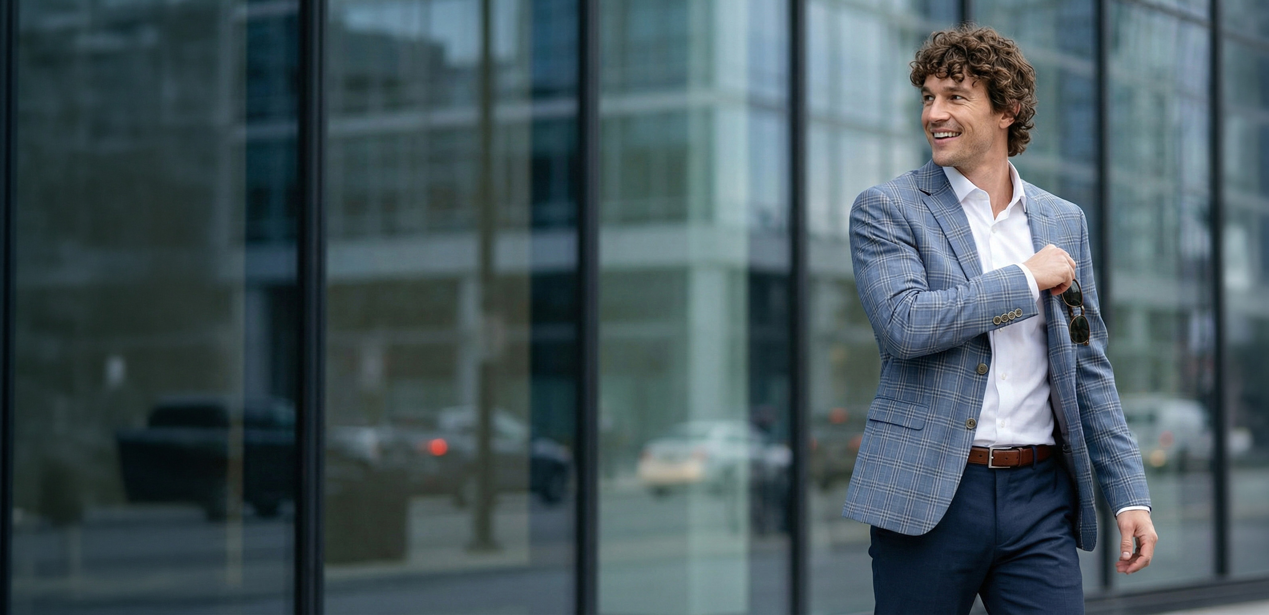 A smiling man with curly hair wearing a blue plaid blazer and a white collared shirt outdoors.
