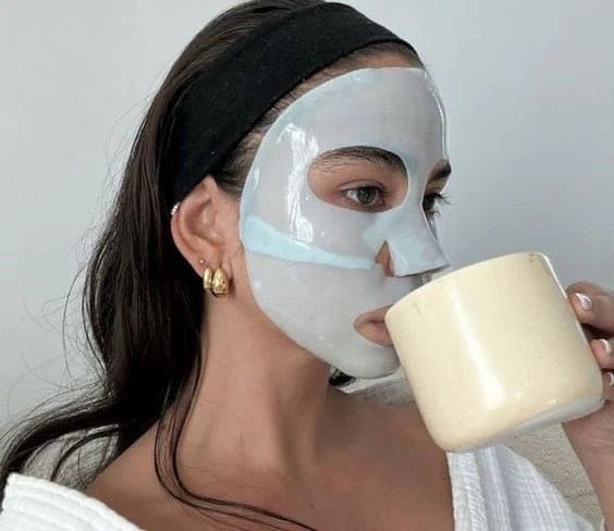 Person wearing a sheet mask, drinking from a beige mug.
