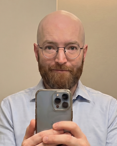 A bald man with a beard and glasses taking a selfie in a mirror with his phone.