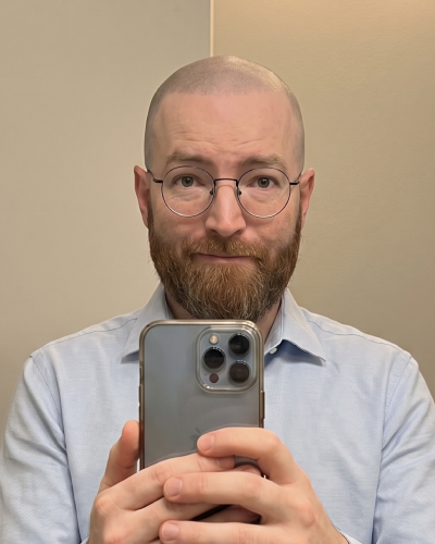 A man with a shaved head, beard, and glasses takes a selfie in a mirror with his phone.