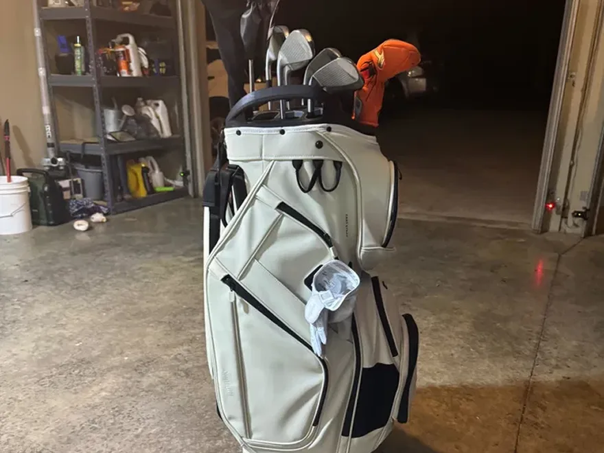 A white and black golf bag full of clubs sits on the concrete floor of a garage.