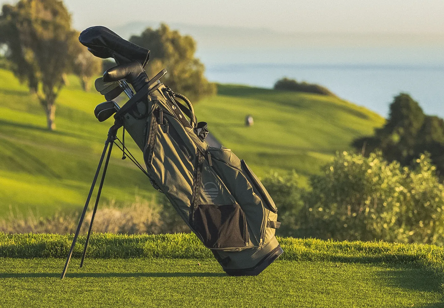 An olive green golf bag with clubs stands on a golf course overlooking the water.