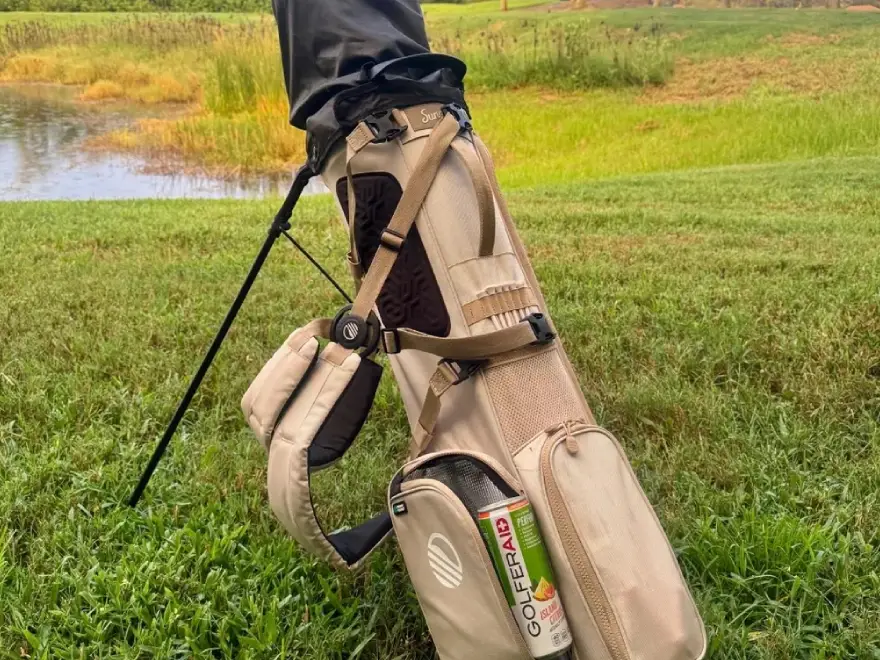 A tan golf stand bag sits on the grass near a pond with a beverage can in its pocket.