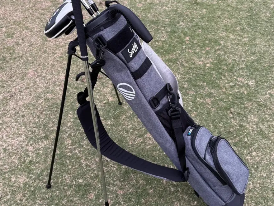 A grey golf bag with clubs stands on a grassy lawn, supported by its built-in stand.