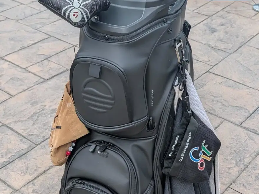 A close-up shot of a black golf bag with accessories, including a spider-themed club cover, on a stone surface.