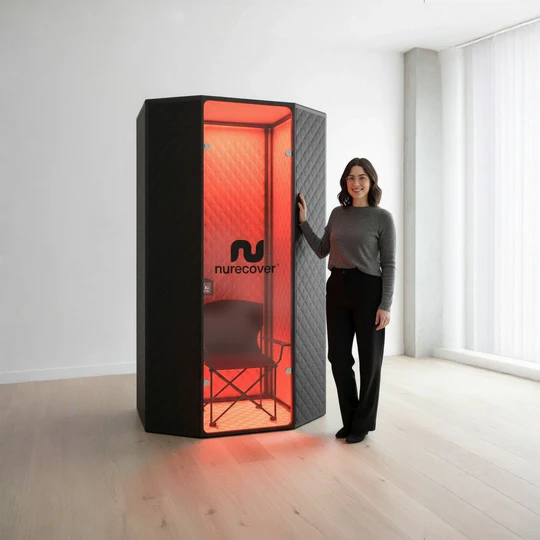 A woman stands next to a tall, hexagonal pod with a red-lit interior containing a chair.