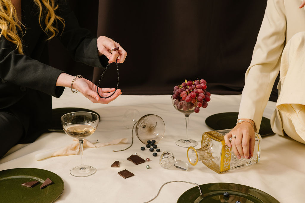 Two people seated with a necklace, drinks, grapes, and chocolate on a table.