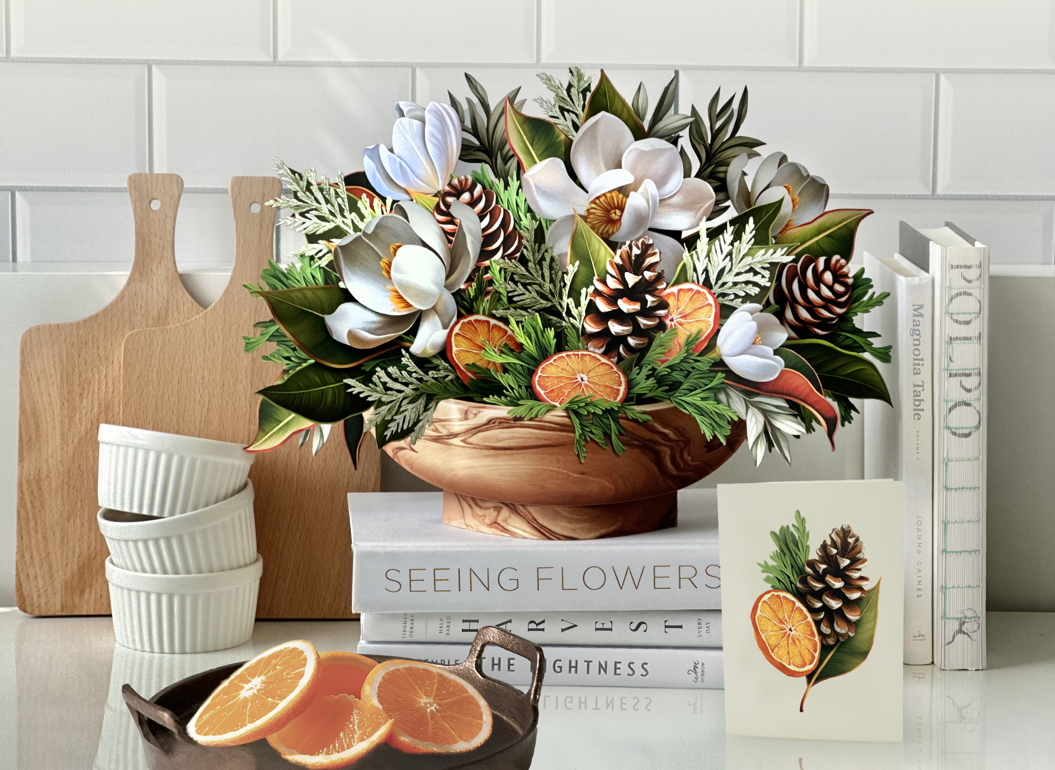 Floral arrangement with pinecones and oranges in a wooden bowl, books stacked nearby.