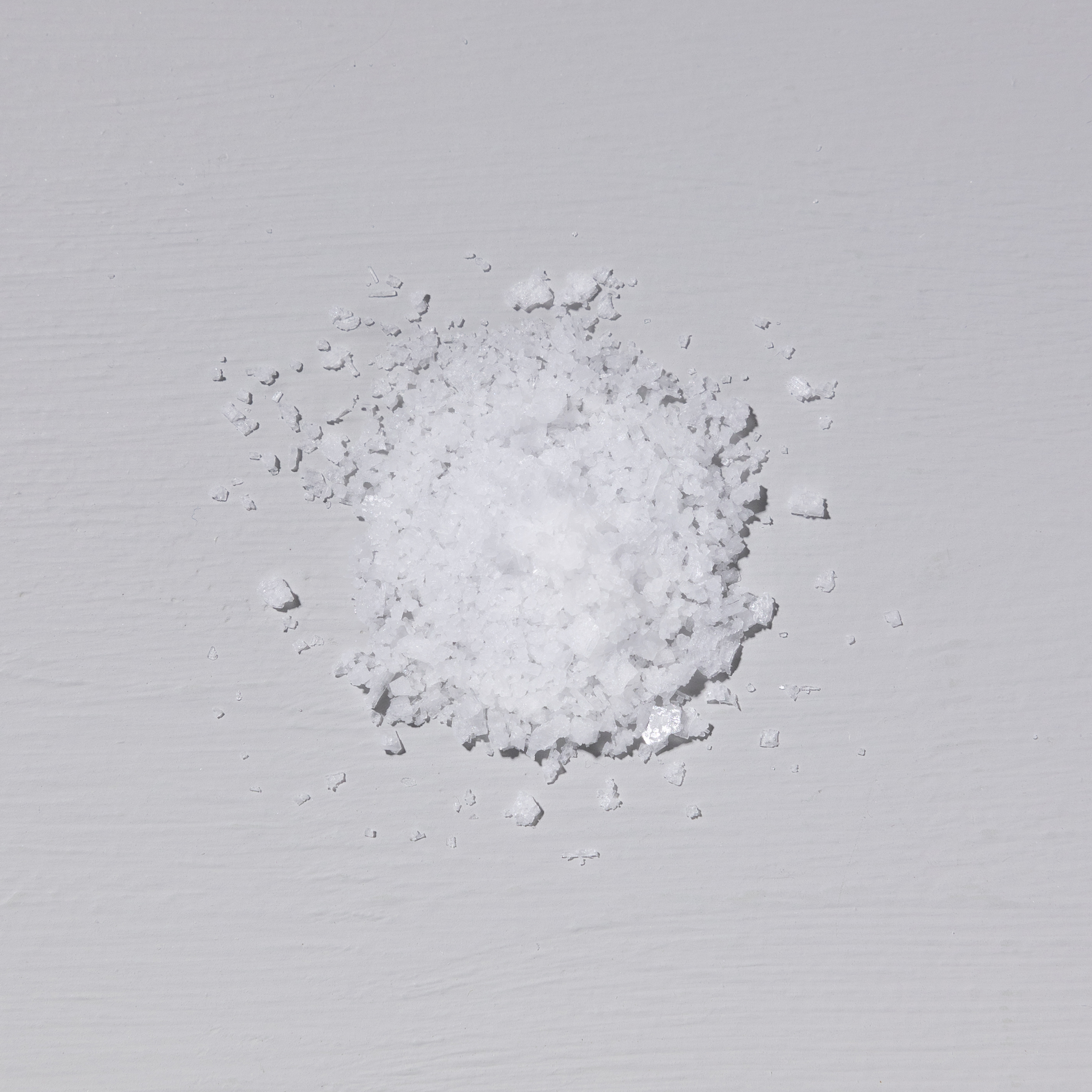 An overhead view of a small pile of coarse white crystals, likely salt, on a light gray surface.
