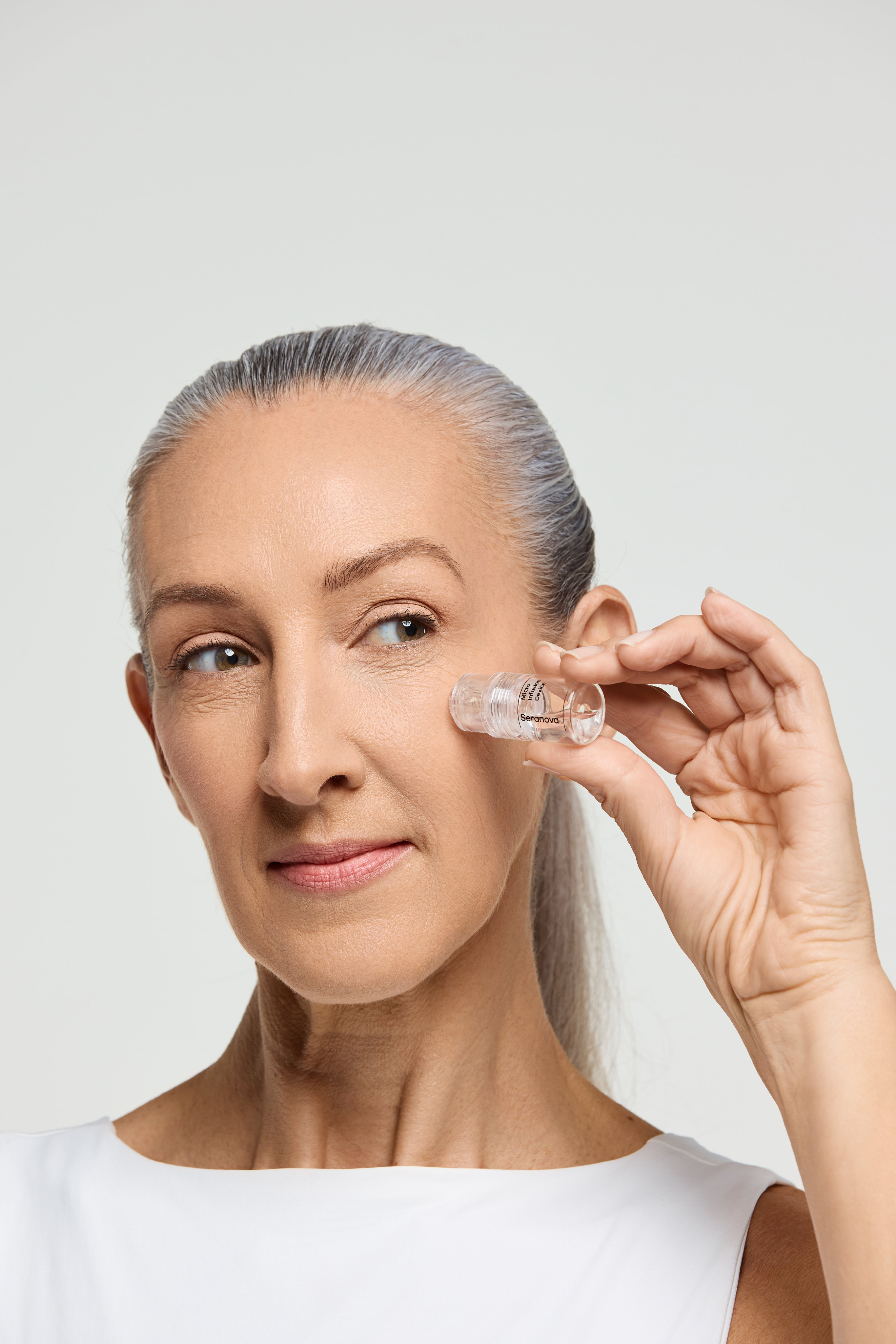 A woman with gray hair holds a small clear vial of serum to her cheekbone against a white background.
