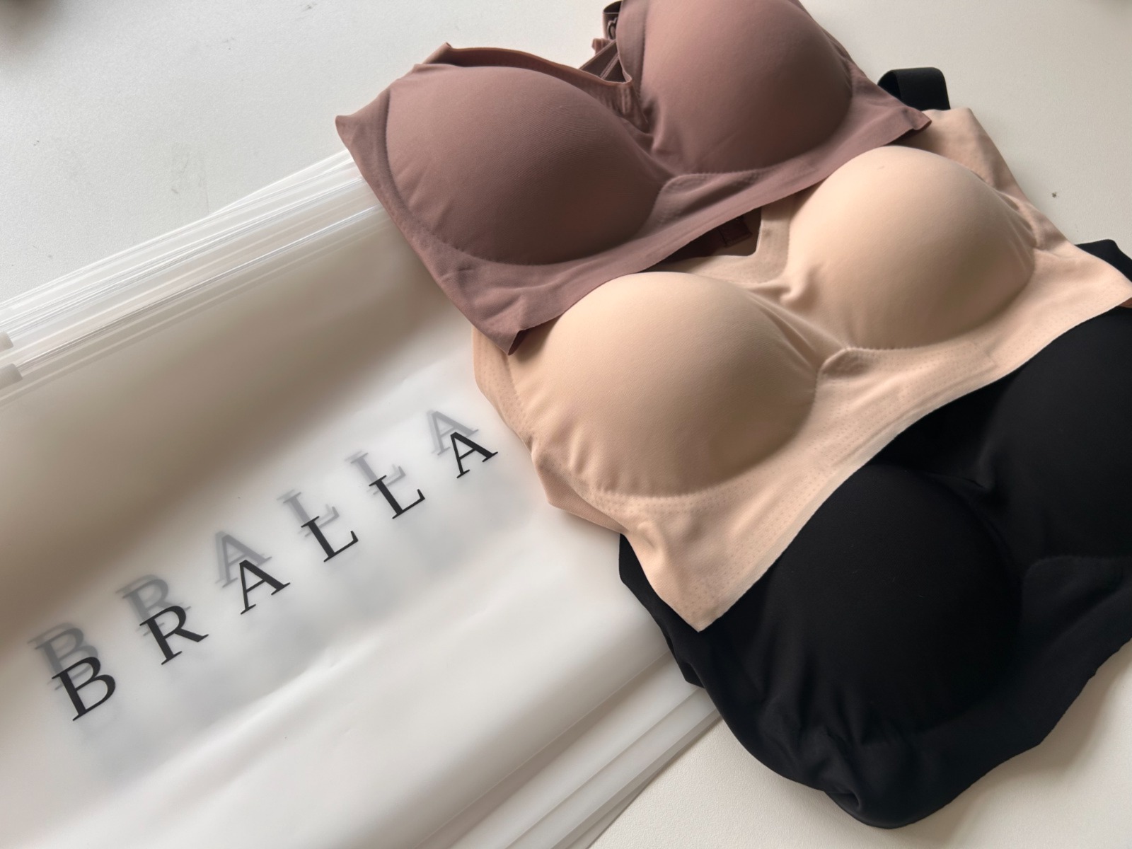 Three seamless bras in neutral tones of brown, beige, and black next to a frosted Bralla bag.