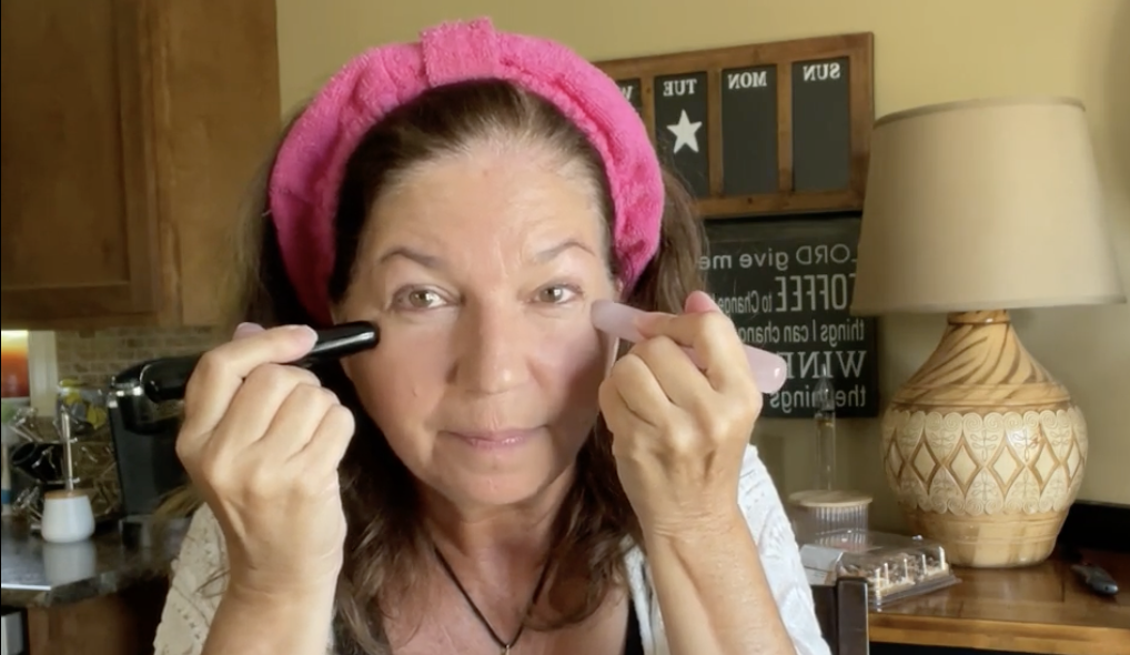 A woman holds two metal tuning fork-like tools to her cheeks while smiling at the camera.