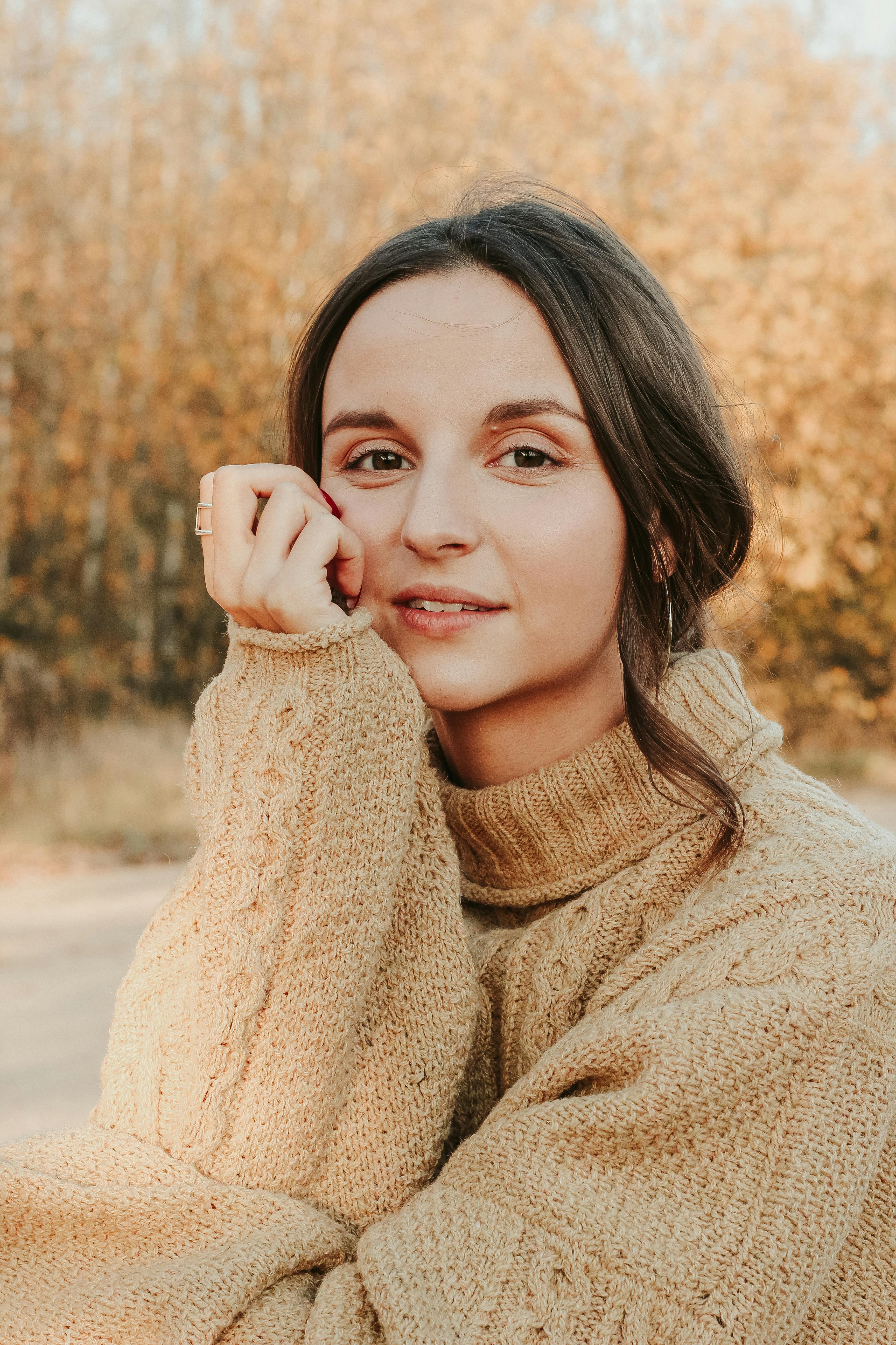 A woman in a tan knit sweater rests her chin on her hand and smiles at the camera.
