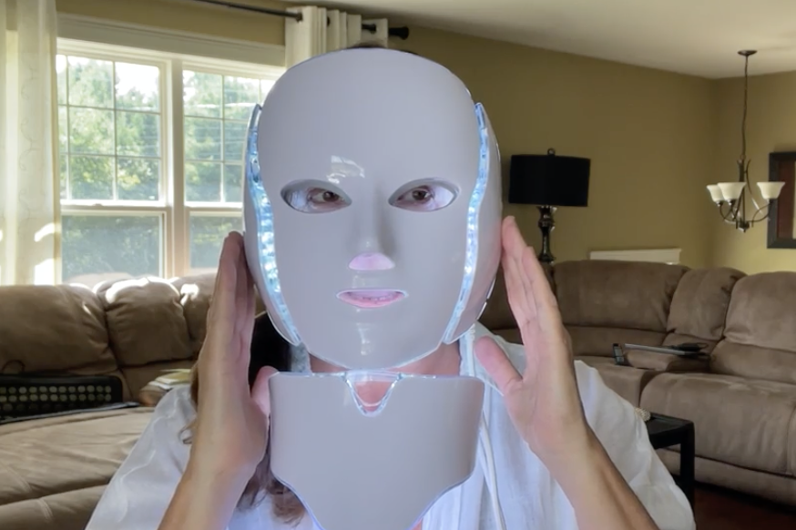 A person holds a white, glowing LED therapy mask and neck attachment up to their face.