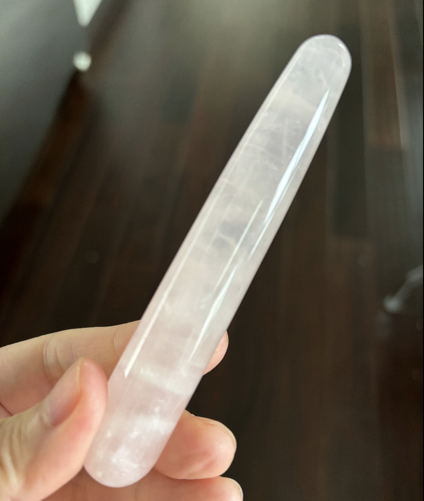 A hand holds a long, smooth, polished rose quartz crystal wand against a dark wood background.