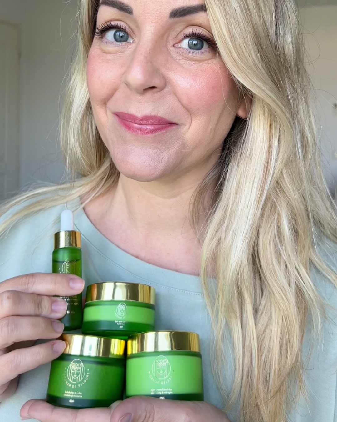 Person holding green skincare products with gold lids, smiling at the camera.