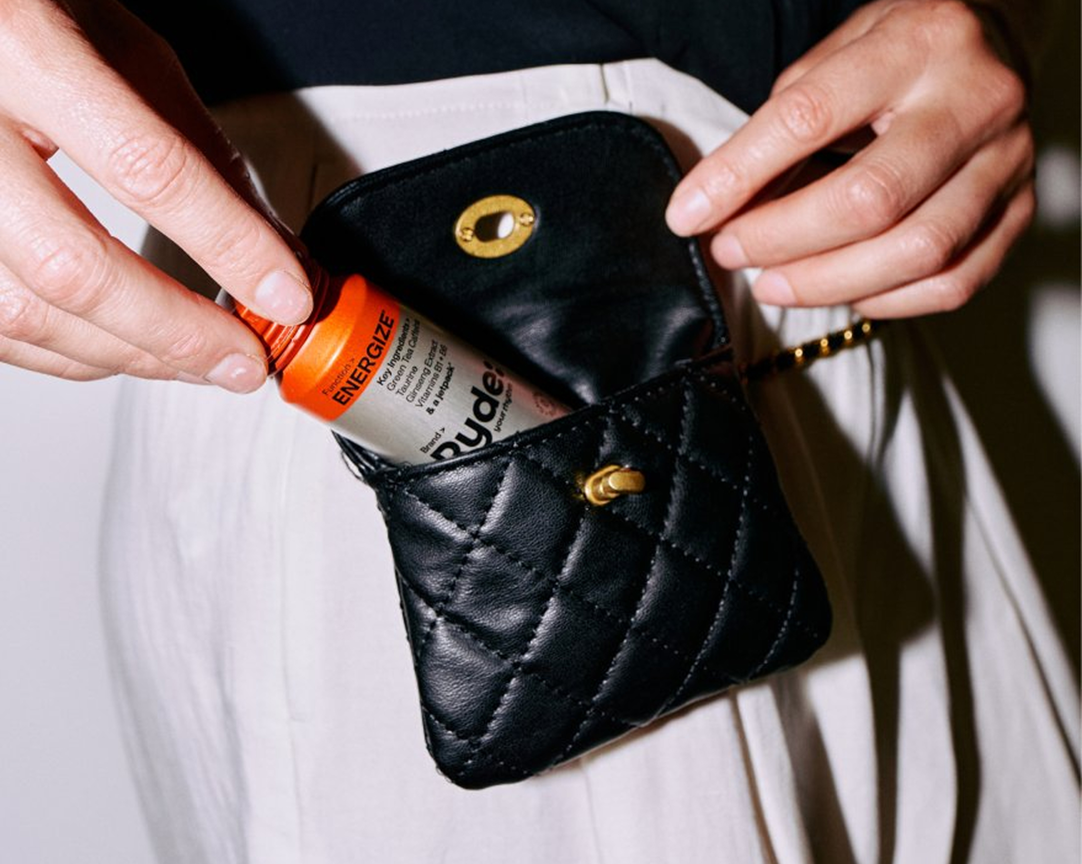 A person puts a bottle labeled 'Energize' into a small, black, quilted handbag.