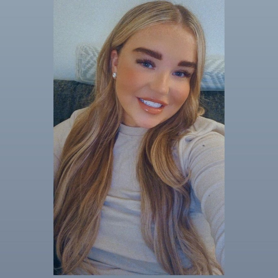 Person smiling with long, wavy blonde hair, wearing a light-colored top.