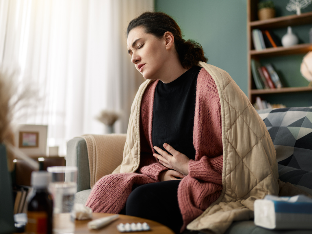 A woman sits on a couch wrapped in a blanket, holding her stomach with a pained expression.