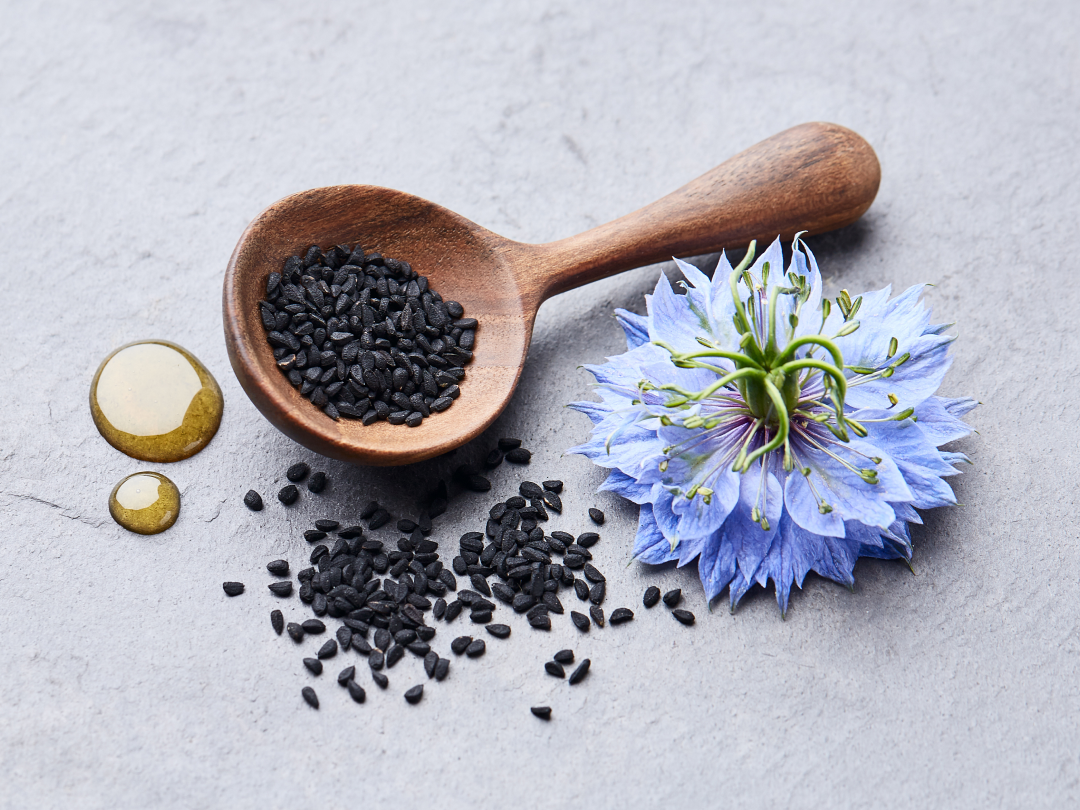 A wooden spoon of black cumin seeds next to a blue flower and drops of oil.
