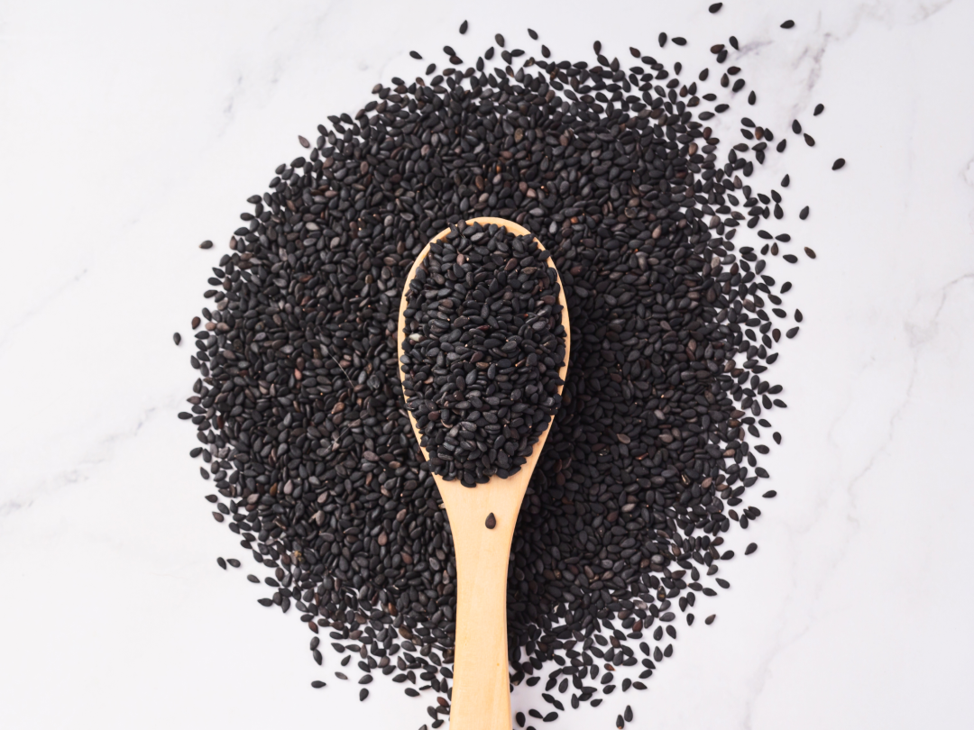An overhead view of a wooden spoon filled with black sesame seeds on a white marble surface.