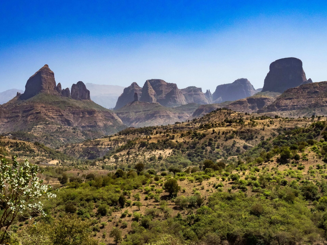 A rugged, rocky mountain range with unique formations rises above a green valley under a clear blue sky.