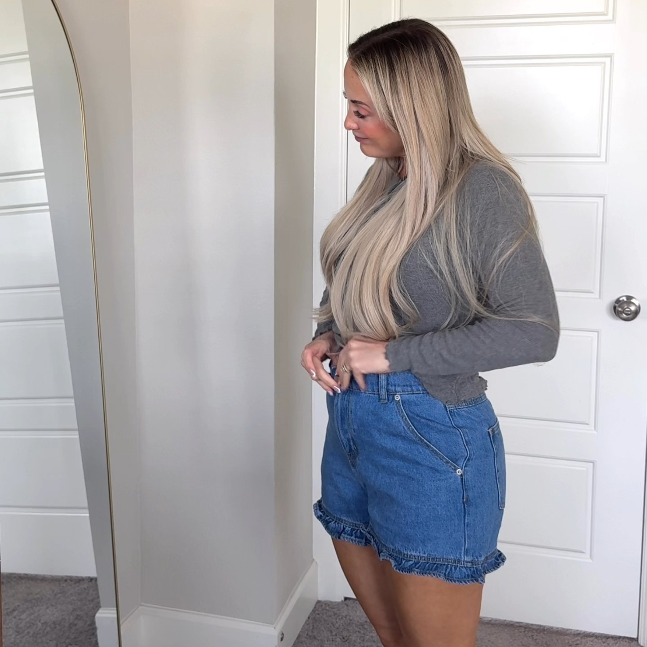 A woman with long blonde hair adjusts her ruffled denim shorts while looking in a mirror.