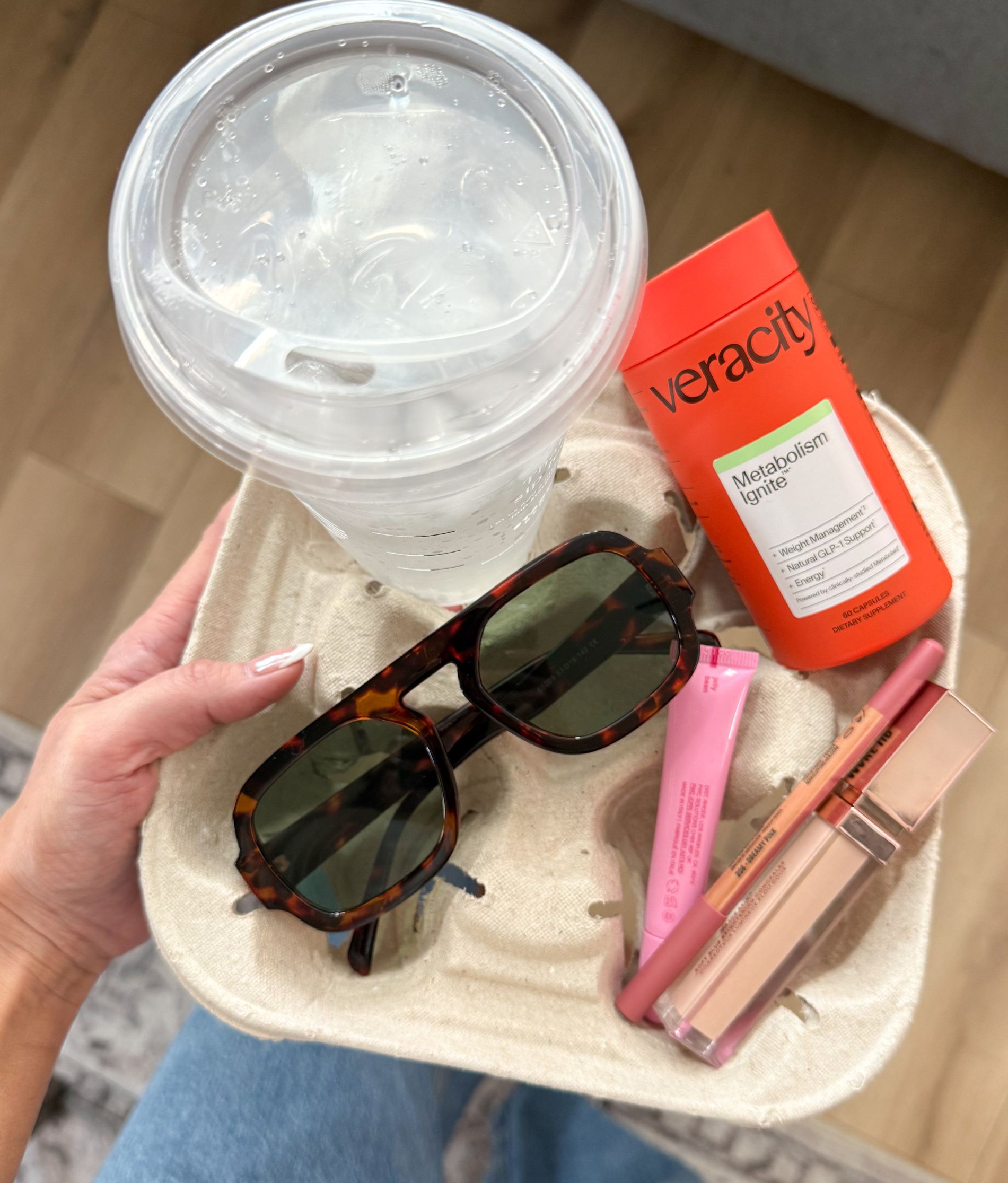 A hand holds a drink carrier with a beverage, supplements, sunglasses, and makeup.