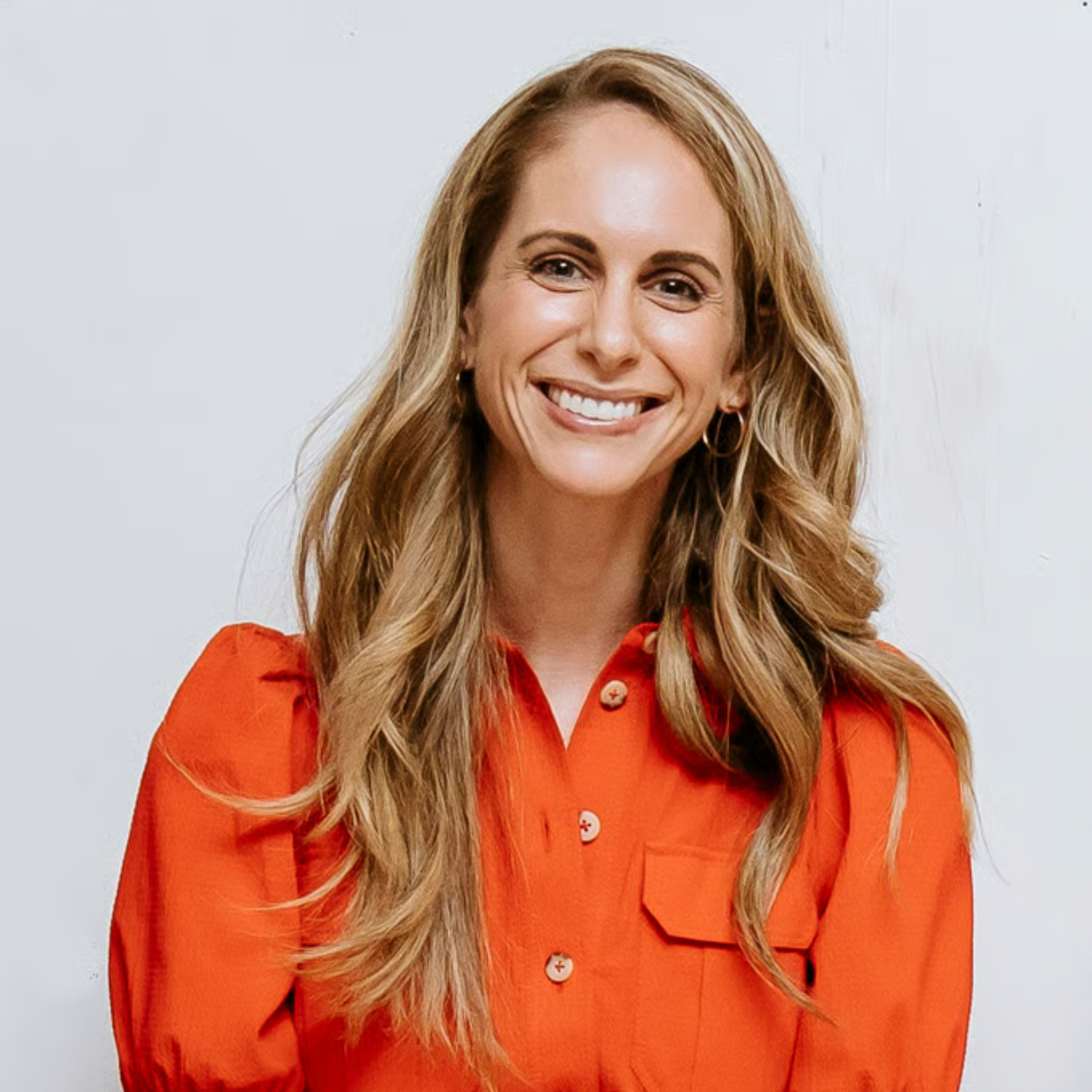 A smiling woman with long, wavy, light brown hair wearing a bright orange button-up shirt.