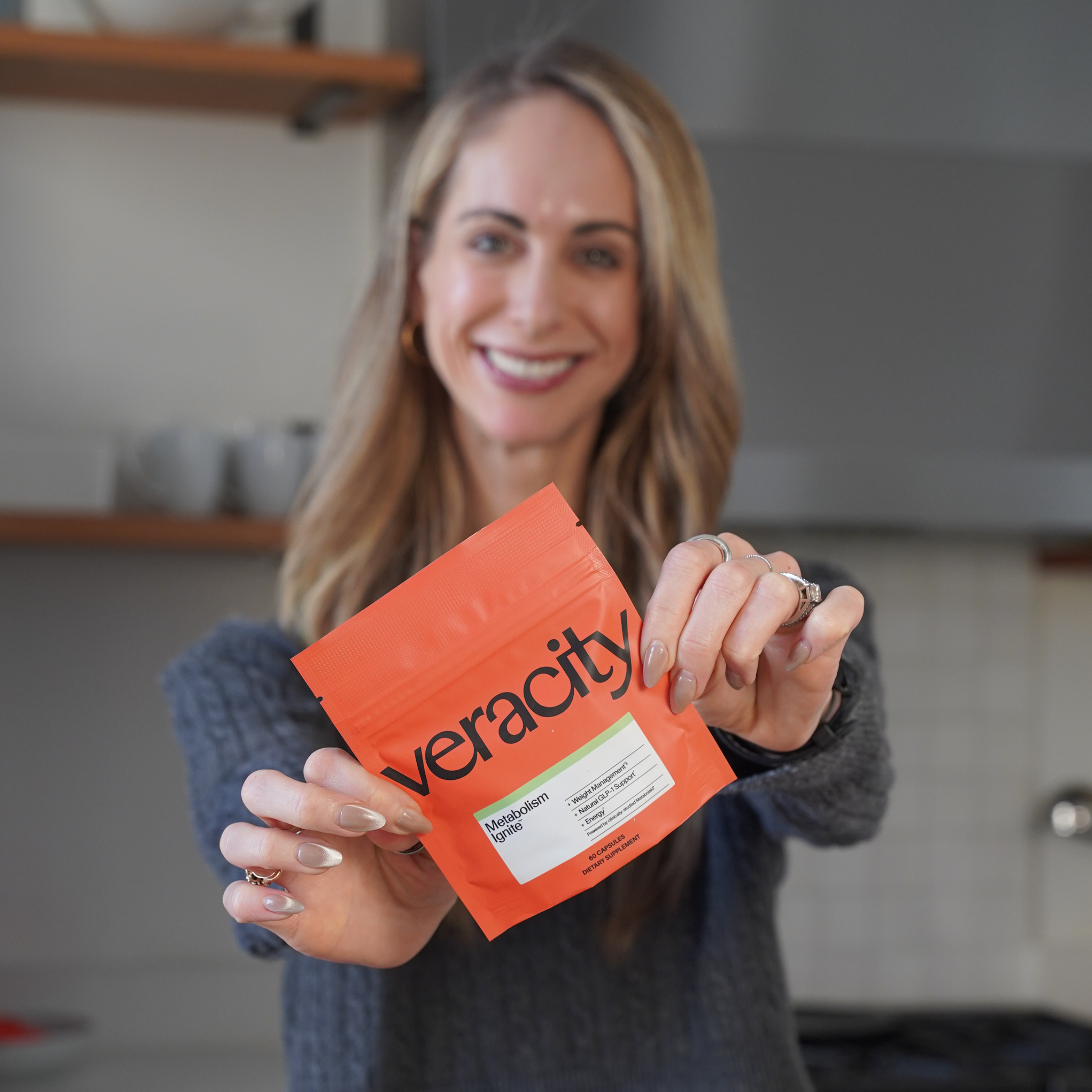 A smiling woman in a grey sweater holds an orange pouch of Veracity Metabolism Ignite toward the camera.