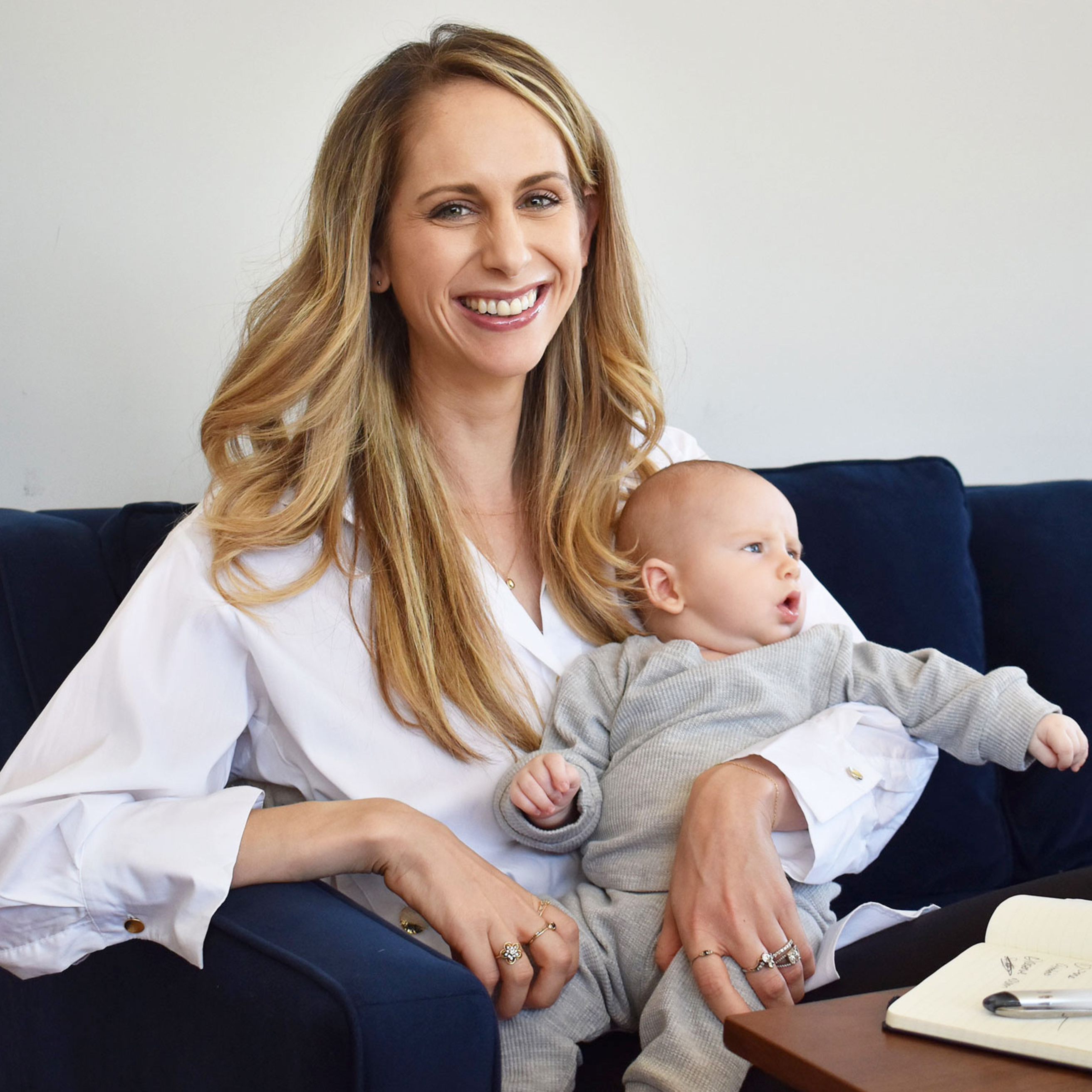 A smiling woman with long blonde hair holds a baby while sitting on a blue couch.