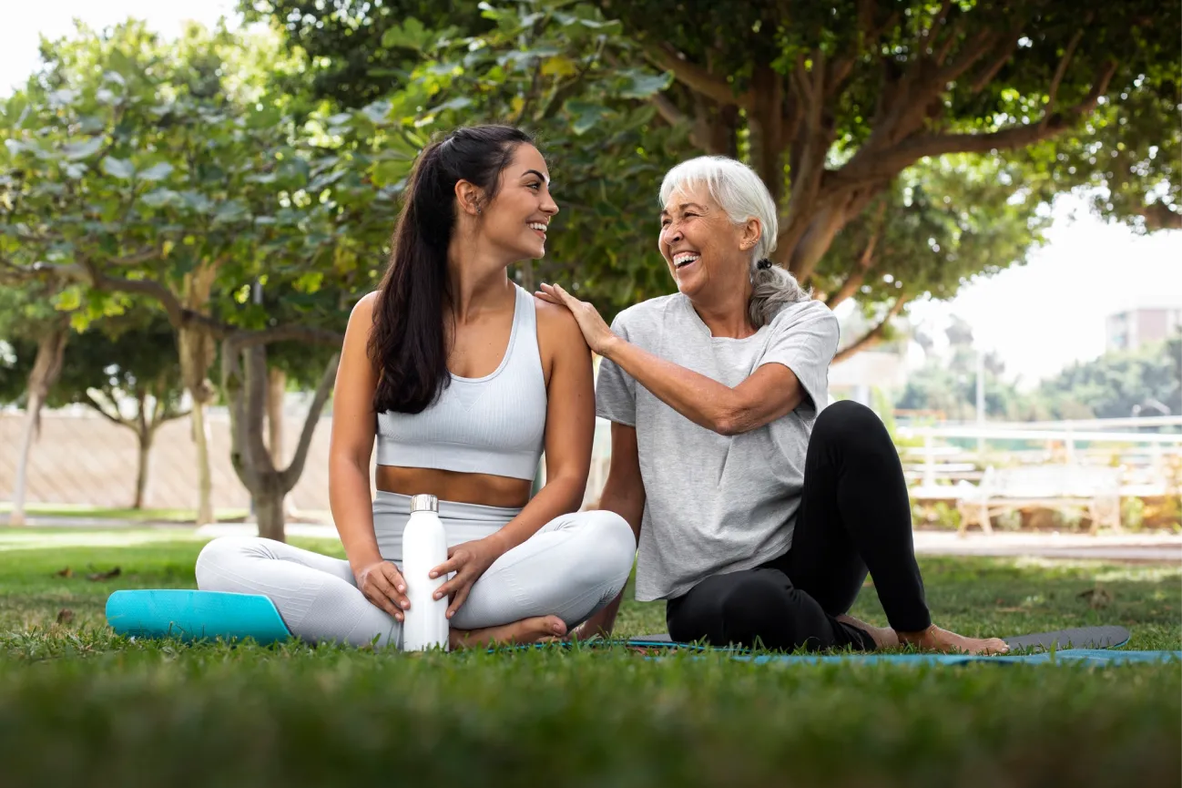 Two women sitting on yoga mats in a park, smiling at each other.