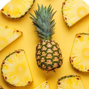 A whole pineapple surrounded by several fresh pineapple slices on a yellow background.